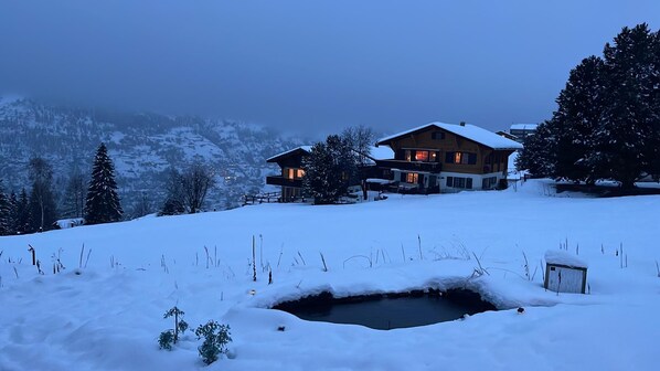 A two-story wooden house with a snow-covered roof, surrounded by snow-covered trees and a mountainous backdrop.