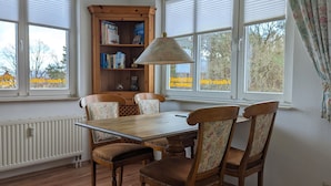 A dining area with a wooden table and chairs, a bookshelf, and large windows with blinds. - Strandnest holiday apartment (Karlshagen)