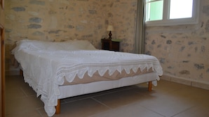 A bed with a white lace cover in a room with stone walls and a window.
