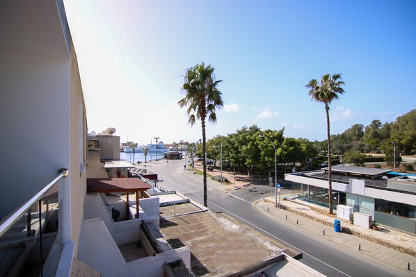 A modern building with a flat roof, a balcony with a metal railing, and a view of a street with palm trees and a body of water in the distance.