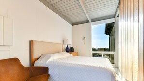 A bedroom with a bed, a chair, a nightstand with a lamp, and a window with a view.