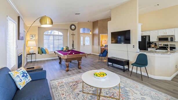 A living room with a pool table, a sofa with a decorative pillow, a round coffee table with fruit, and a kitchen area with a TV and appliances. - New! Cozy Modern Pool Home, Close to Downtown and The Strip (North Las Vegas)