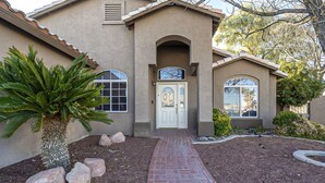 A stucco house with a brick pathway leading to a white door, surrounded by landscaping and a palm tree. - New! Cozy Modern Pool Home, Close to Downtown and The Strip (North Las Vegas)