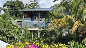 A house with a covered porch, surrounded by lush tropical vegetation and a variety of colorful flowers. - VILLA PAMEYAQUE T3 SUPERB SEA VIEW AND SWIMMING POOL (Rivière-Salée)