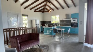 A spacious living area with a sofa, a dining table, and chairs. A kitchen with blue cabinets and modern appliances is visible in the background. - VILLA PAMEYAQUE T3 SUPERB SEA VIEW AND SWIMMING POOL (Rivière-Salée)