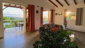 A spacious living area with a large plant, a sofa, and a dining table. There is a view of a balcony and a door leading outside. - VILLA PAMEYAQUE T3 SUPERB SEA VIEW AND SWIMMING POOL (Rivière-Salée)
