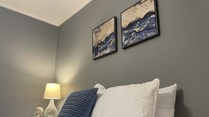 Ein Schlafzimmer mit einem Bett, einem Nachttisch mit Lampe und zwei gerahmten Kunstwerken an der Wand.