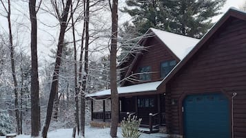 A log cabin with a covered porch and a garage, surrounded by snow-covered trees.