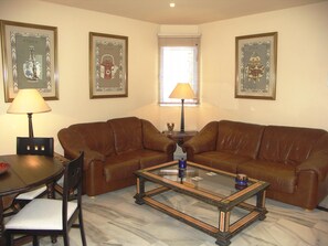 Un salon avec deux canapés en cuir marron, une table basse en verre, une table à manger avec des chaises et des œuvres d’art encadrées accrochées aux murs.