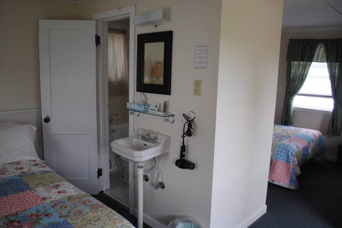 Un dormitorio con cama, una puerta blanca, un baño con lavamanos y espejo, y una ventana con cortinas.