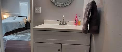 Baño con lavamanos, espejo y perchero. Se ve una cama en la habitación contigua.