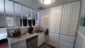 Una cocina compacta con gabinetes blancos, lavavajillas integrado, cafetera y ventana con persianas.