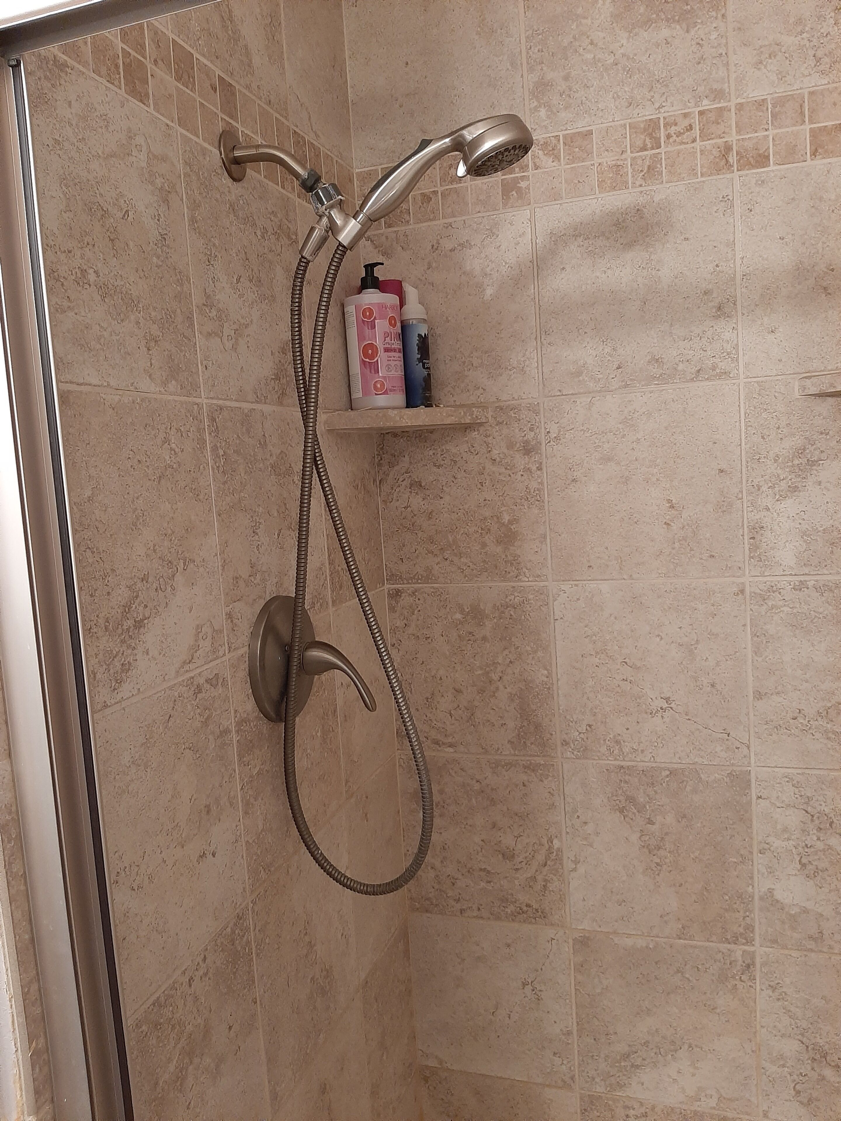A shower with a handheld showerhead and a flexible hose, a shelf with toiletries, and a tiled wall.