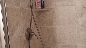 A shower with a handheld showerhead and a flexible hose, a shelf with toiletries, and a tiled wall.