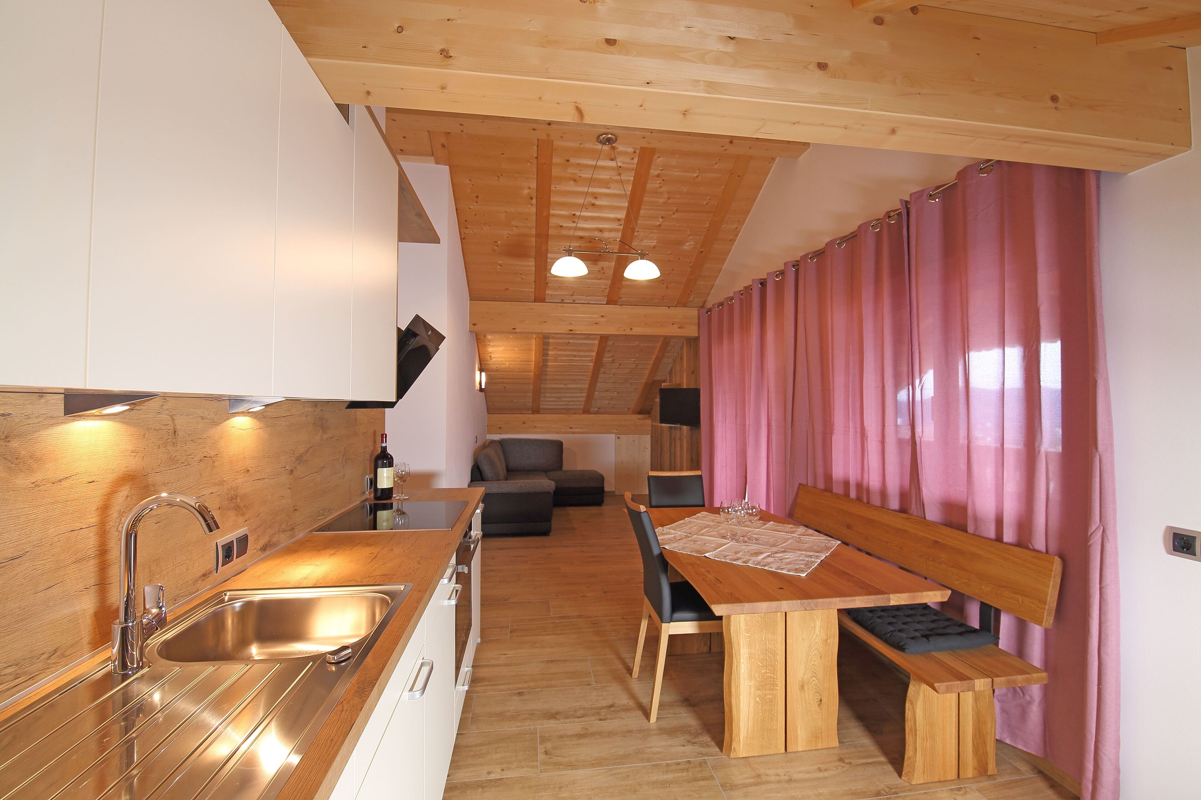 Eine moderne Küche mit Holzarbeitsplatte und Edelstahlspüle. Es gibt einen Essbereich mit Holztisch und Bank. Der Raum verfügt über Holzbalken an der Decke und rosa Vorhänge.