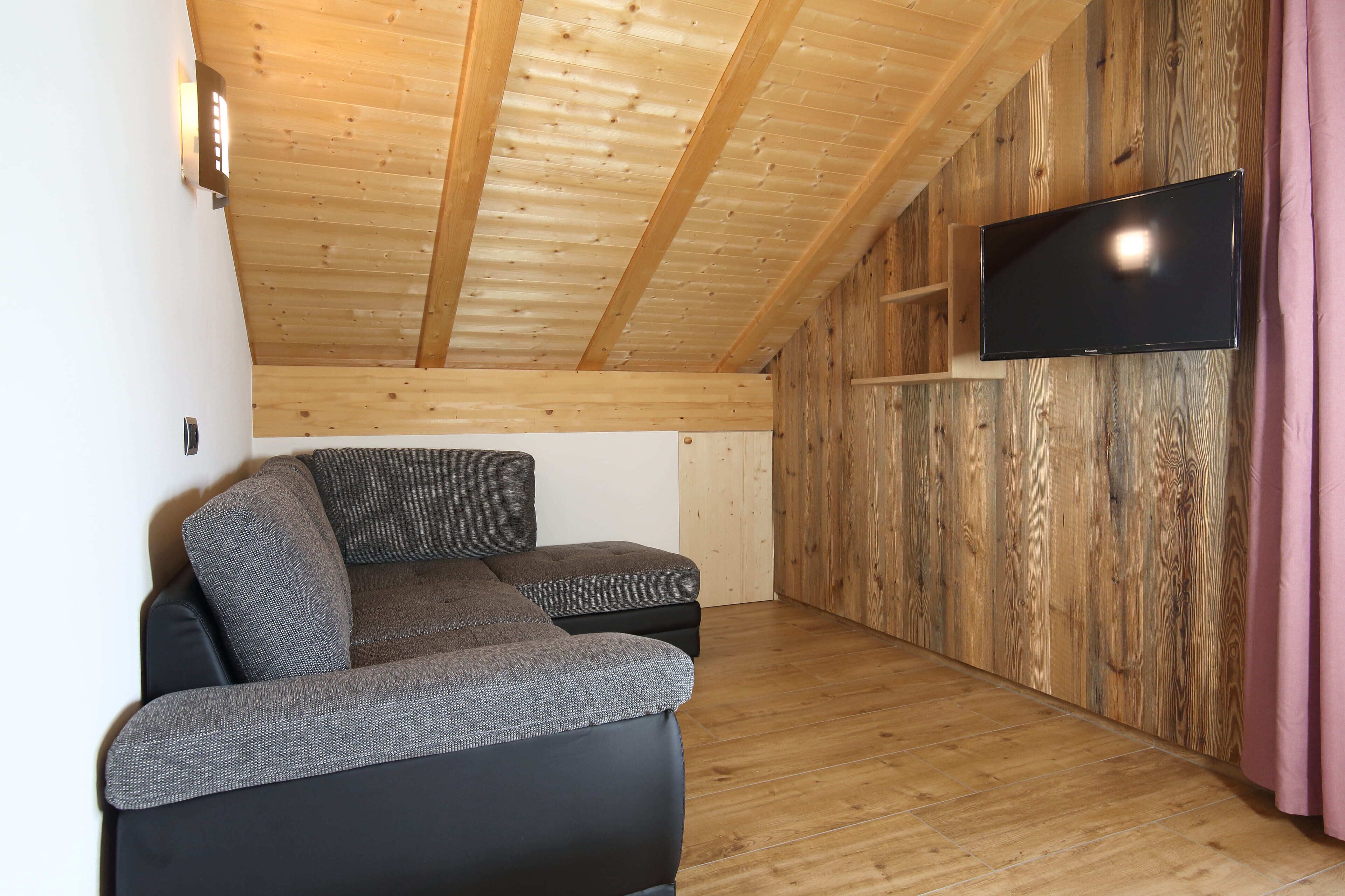 Ein moderner, kompakter Wohnraum mit Holzboden und -wänden, einem an der Wand befestigten Flachbildfernseher und einem Ecksofa.