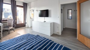 Habitación de hotel con una cama grande, un televisor sobre un tocador blanco, una silla y vistas al exterior.