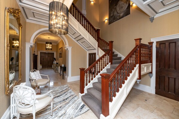 Una imponente escalera de madera con barandillas y un candelabro, que conduce a un descansillo con un espejo grande y zona de estar.