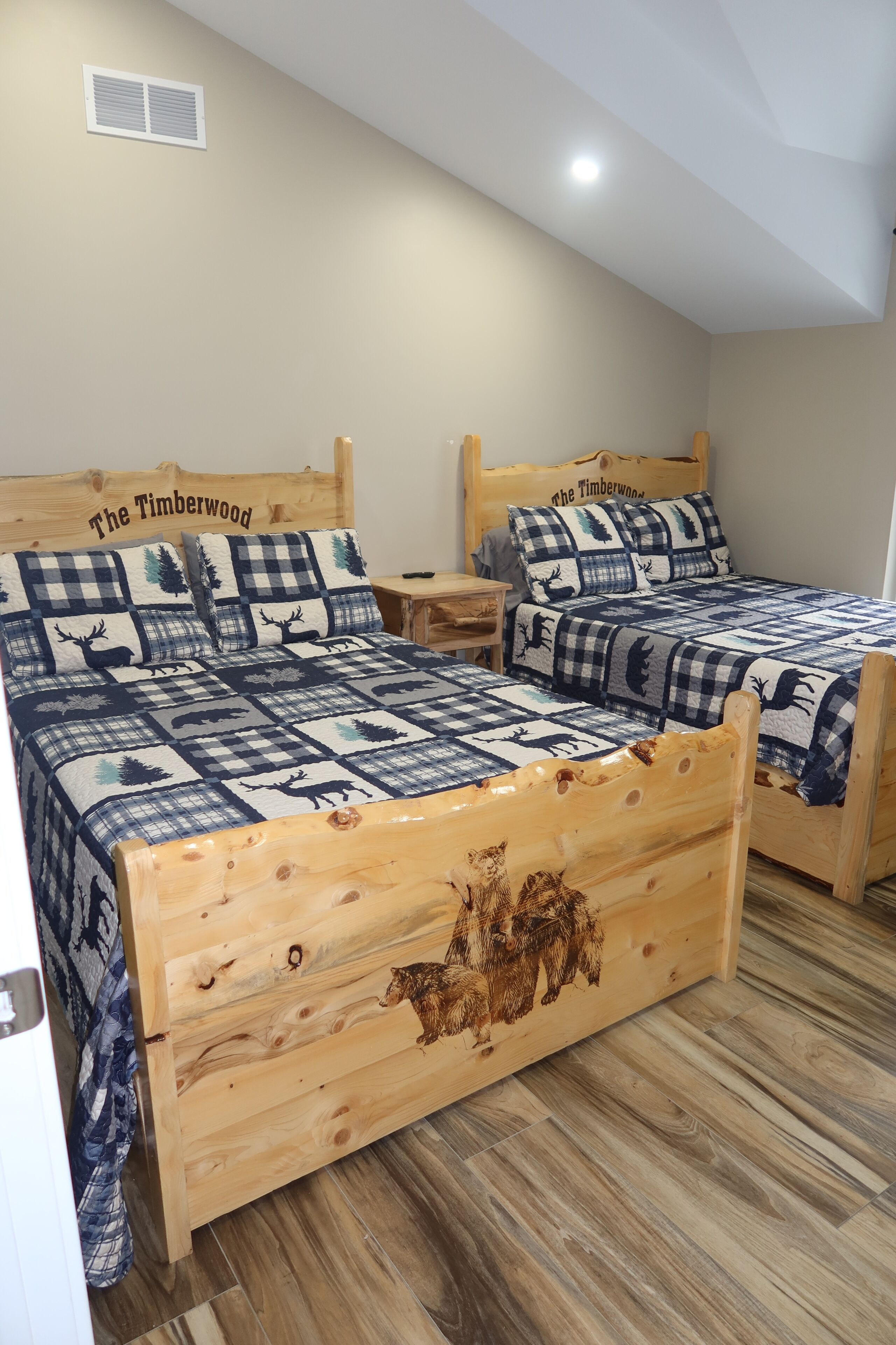 Deux lits en bois, dotés de draps à carreaux et d’une tête de lit décorée sur le thème de la forêt, dans une pièce au parquet en bois et à l’aménagement simple et fonctionnel.