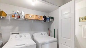 Uma lavanderia com duas máquinas de lavar brancas, prateleiras com produtos de limpeza e uma porta com uma toalha pendurada.