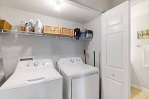 A laundry room with two white washing machines, shelves with cleaning supplies, and a door with a towel hanging on it. - Topsail at Schooner Court (St. Augustine Beach)