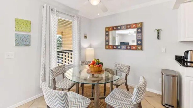 Uma área de refeições com mesa de vidro rodeada por cadeiras estampadas, um ventilador de teto, um espelho decorativo e uma pequena lixeira.