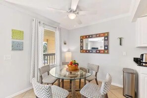 A dining area with a glass table surrounded by patterned chairs, a ceiling fan, a decorative mirror, and a small trash bin.
