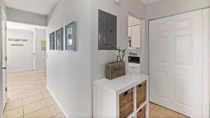 Corredor com porta branca, um console branco com cestos e uma cozinha ao fundo.