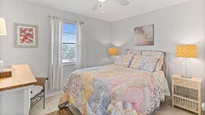 Quarto com cama, escrivaninha, cadeira, mesa de cabeceira, berço, ventilador de teto e janela com cortinas.