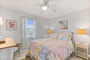 A bedroom with a bed, a desk, a chair, a nightstand, a crib, a ceiling fan, and a window with curtains. - Topsail at Schooner Court (St. Augustine Beach)
