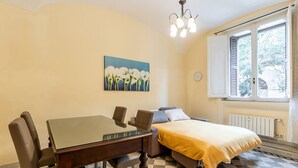 A bedroom with a bed, a table, and chairs. There is a window with shutters and a painting on the wall.
