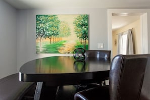 Un coin repas avec une table ronde, des chaises et un tableau représentant un chemin bordé d’arbres accroché au mur.