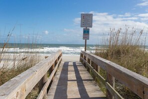 A wooden boardwalk leading to the beach with signs indicating beach access and a lifeguard station.