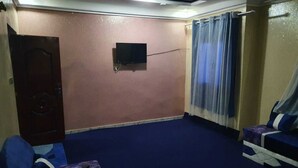 A room with a dark blue carpet, a television mounted on the wall, a door, and two blue sofas with cushions. - Corporate Apartment (Nouakchott)