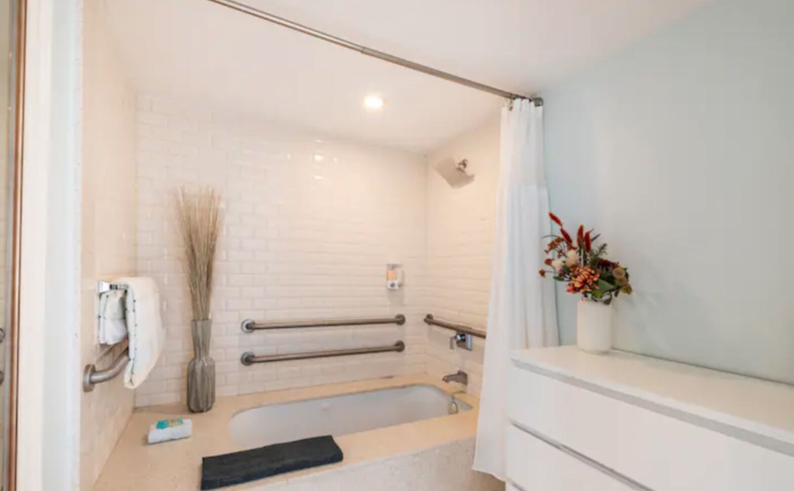 A modern bathroom with a bathtub, a shower, and a toilet. There are handrails for accessibility, a towel rack, and a vase with flowers on the counter.