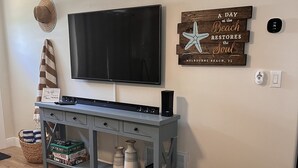 A living room with a flat-screen TV mounted on the wall, a grey entertainment unit with a basket and storage boxes, a woven pouf, and a motivational sign.