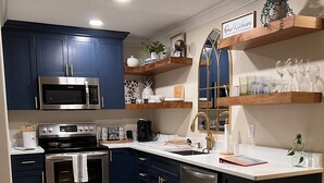 A modern kitchen with dark blue cabinets, stainless steel appliances, wooden shelves, and a decorative rug.