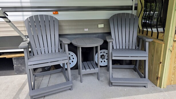 Een klein buitgedeelte met twee grijze Adirondack-stoelen en een ronde tafel, gelegen voor een witte camper met een schuifdeur.