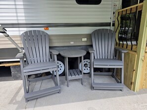 A small outdoor seating area with two grey Adirondack chairs and a round table, positioned in front of a white RV with a sliding door. - Miss Daisy's Place - quiet and relaxing RV camper (Gatlinburg)