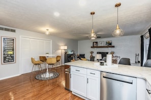 A modern kitchen with white cabinets, stainless steel appliances, and a marble countertop. There is a dining area with a round table and chairs, and a living area with a sofa and a fireplace.
