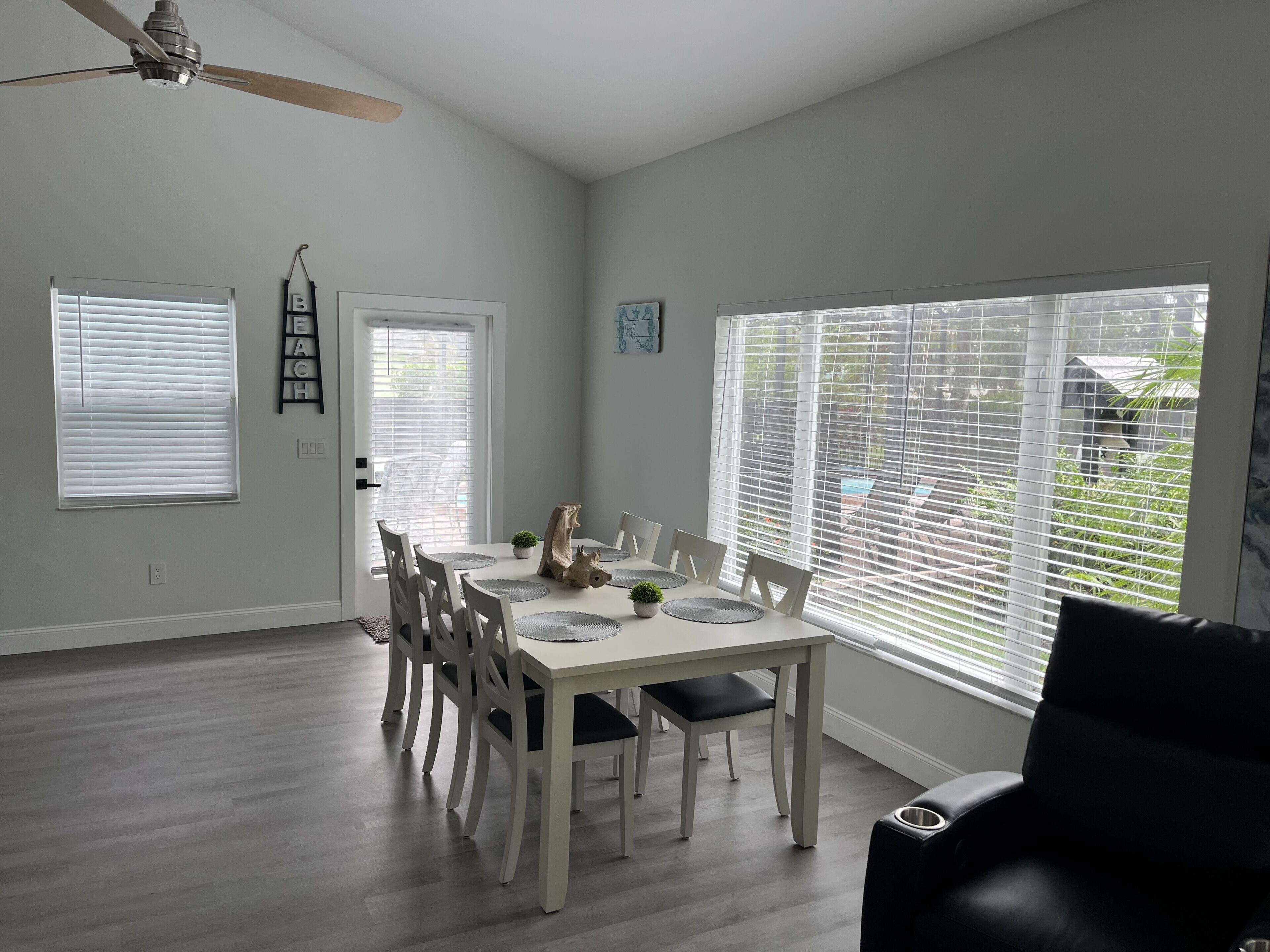 A dining area with a white table and chairs, a ceiling fan, and large windows with blinds.