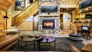 A log cabin interior with a stone fireplace, wooden staircase, and a television mounted on the wall. - 5BR/5BA Cabin, Sleeps 14, Hot Tub/Game Rm, Pool Close, 5min to G'burg/Nat'l Park (Gatlinburg)