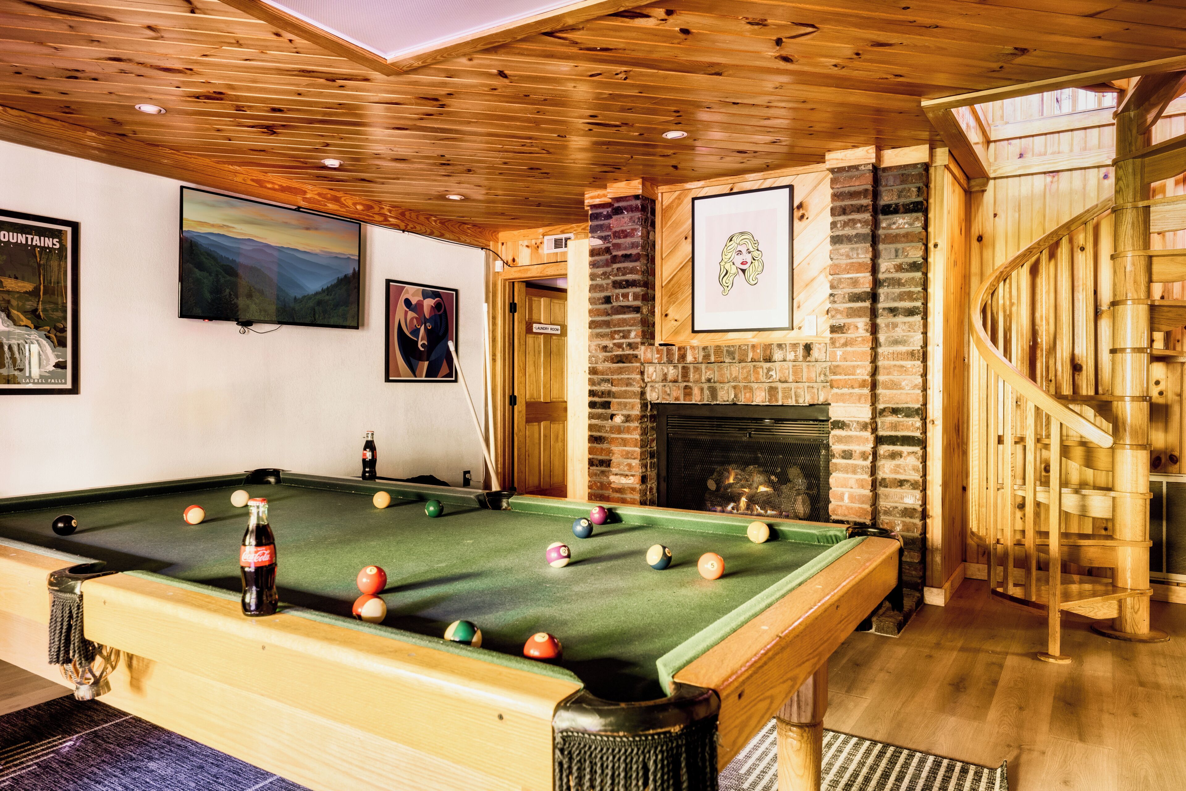 A pool table with balls, a bottle of Coca-Cola, and a fireplace in a room with wooden ceilings and walls.
