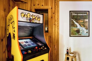 Une borne d’arcade avec le jeu Pac-Man, une affiche des Great Smoky Mountains et un tabouret avec une bouteille et un bol.