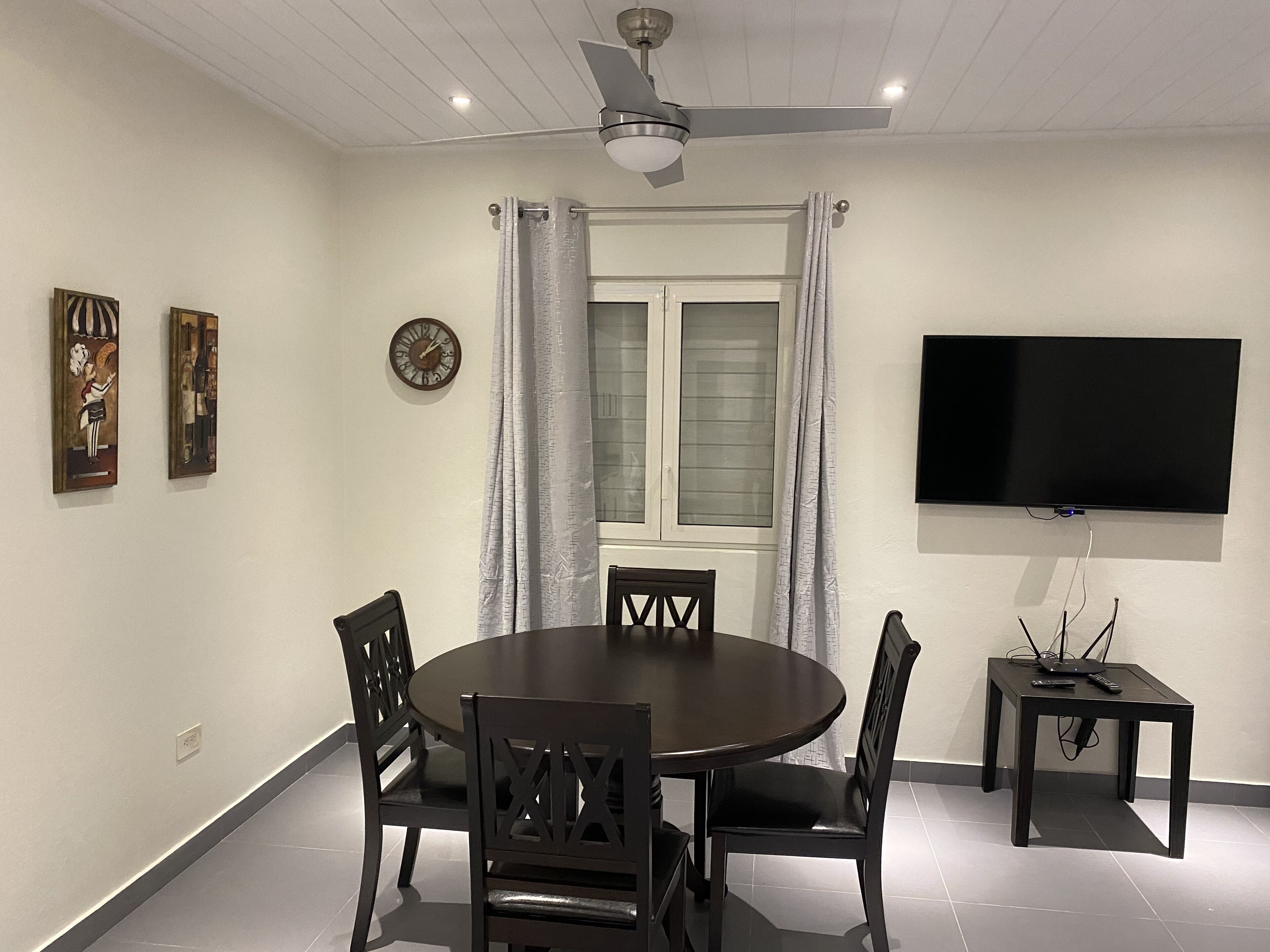 A dining area with a round table, four chairs, a ceiling fan, a TV mounted on the wall, and a window with curtains.