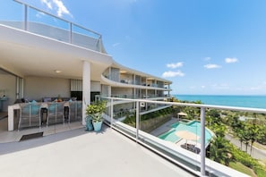 A modern hotel balcony with a pool, seating area, and ocean view.