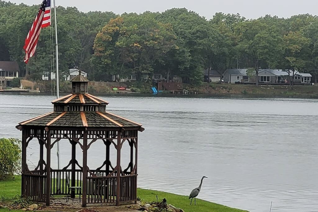 A gazebo by a lake with an American flag, surrounded by houses and trees.