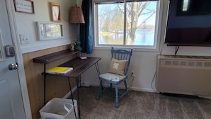 A room with a wooden desk, a blue chair, a window with a view of a lake, a television, and a wall-mounted air conditioner. - Lakeside Quonset Hut, Cozy And Romantic (Paw Paw)