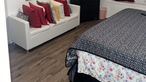 Quarto com sofá branco, cama com colcha florida, televisão de tela plana, espelho e janela com cortinas.