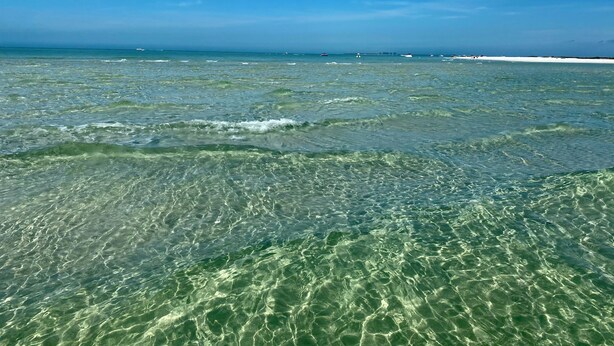 On aperçoit clairement une plage avec une eau peu profonde et des bateaux au loin.